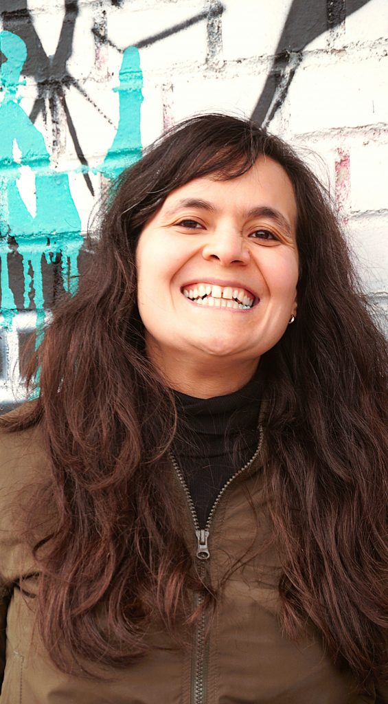 Malika Guellil lächelt und steht vor einem Urban Art Hintergrund. Sie hat dunkle braune lange Haare und braune Augen.