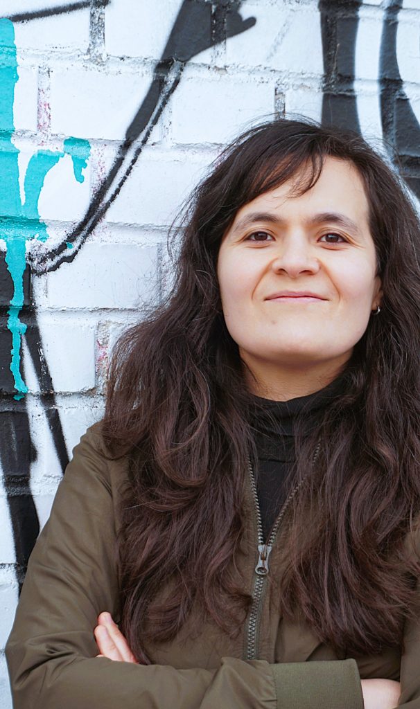 Malika Guellil steht vor einem Urban Art hintegrund. Sie hat die Arme verschrenkt und trägt eine Olivegrüne Jacke. Sie hat dunkelbraune lange Haare und braune Augen.