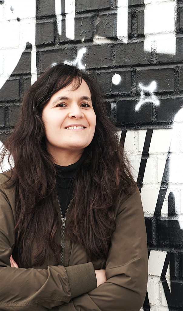 Malika Guellil steht vor einem urban-art Hintergrund. Sie hat dunkelbraune lange Haare und braune Augen. Sie lächelt und hat die Arme unter der Brust verschränkt. Sie trägt eine olive-grüne Weste.